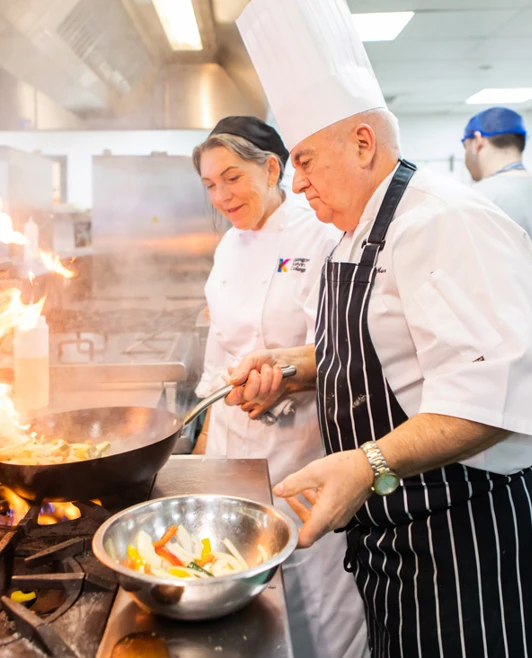 A professional chef in a white uniform and tall hat demonstrates a flambé technique, creating large flames while cooking vegetables in a wok, with a female student watching. A professional chef in a white uniform and tall hat demonstrates a flambé technique, creating large flames while cooking vegetables in a wok, with a female student watching.